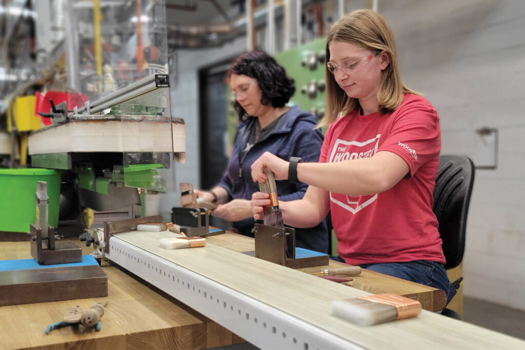 Two female employees working side by side indoors making brushes and wearing safety goggles 