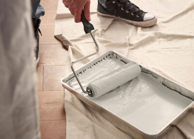 a paint roller on a frame is rolled into a tray of white paint on a covered floor